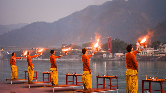 Priests performing the Ganga Aarti ceremony at dusk on the banks of the Ganges River in Rishikesh, holding large brass lamps with flames.