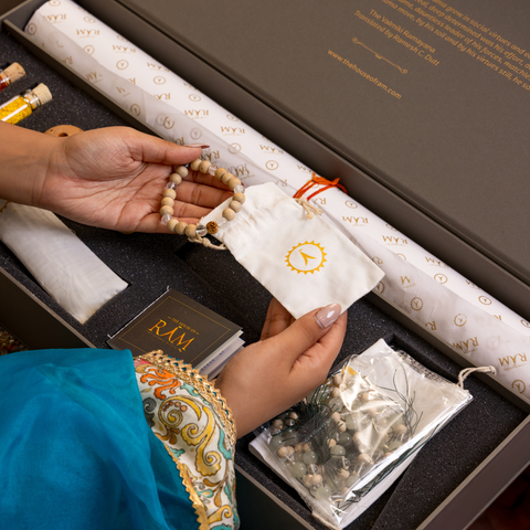 Hands hold a beaded bracelet over a decorative box with a pouch and book labeled "RAM" Elegantly patterned blue cloth adds vibrancy. Hands hold a beaded bracelet over a decorative box with a pouch and book labeled "RAM" Elegantly patterned blue cloth adds vibrancy.