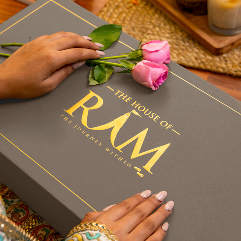 Hands gently hold pink roses atop a dark grey box labeled "The House of Rām, The Journey Within" in gold. The scene conveys elegance and warmth. Hands gently hold pink roses atop a dark grey box labeled "The House of Rām, The Journey Within" in gold. The scene conveys elegance and warmth.