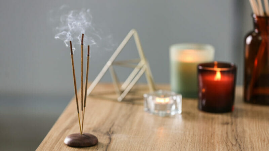 Three smoking incense sticks in a wooden holder on a table with lit candles and a decorative geometric object in the background.