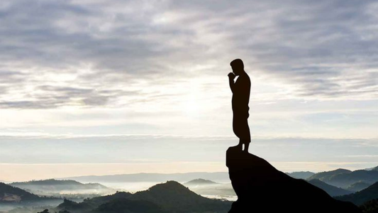 A silhouette of a person standing on a rocky outcrop, contemplatively gazing over misty hills under a cloudy sky.