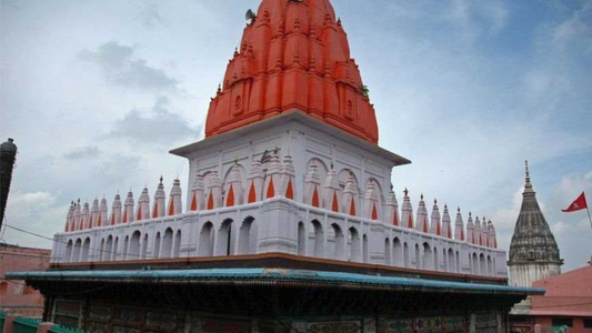 A multi-tiered Hindu temple with an orange domed top, surrounded by ornate white architecture and a cloudy sky backdrop.