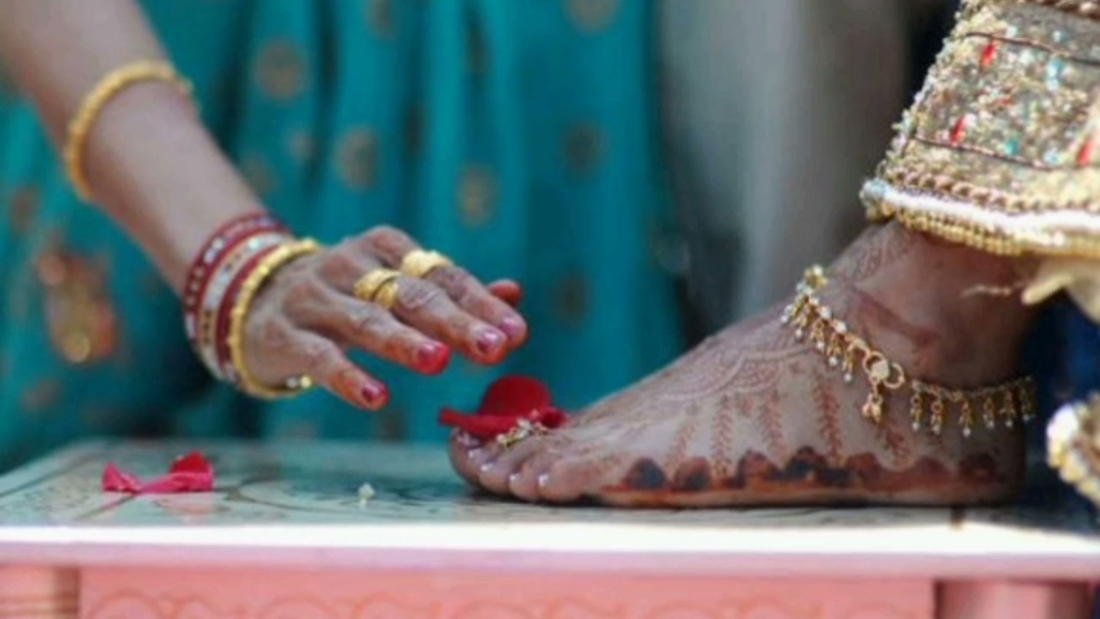 A hand adorned with colorful bangles touches a henna-decorated foot, symbolizing reverence. Rose petals lie nearby, enhancing the ceremonial feel.