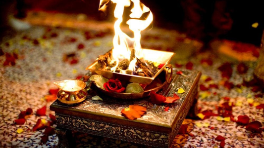 A decorative altar with a fire in a metal bowl, surrounded by rose petals and small oil lamps on an ornate surface.
