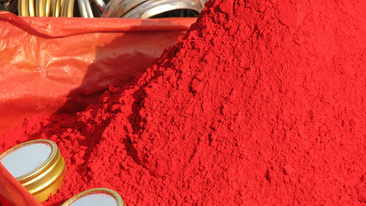 A mound of vibrant red powder is displayed on a red plastic sheet. A few small metallic lids are partially visible, adding a touch of contrast.