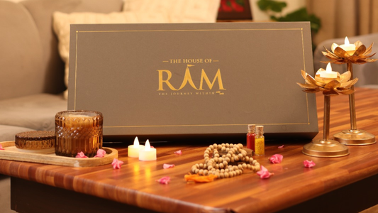 A decorative arrangement featuring "The House of RĀM" box, candles, colorful powders, a wooden tray, and prayer beads on a wooden table.