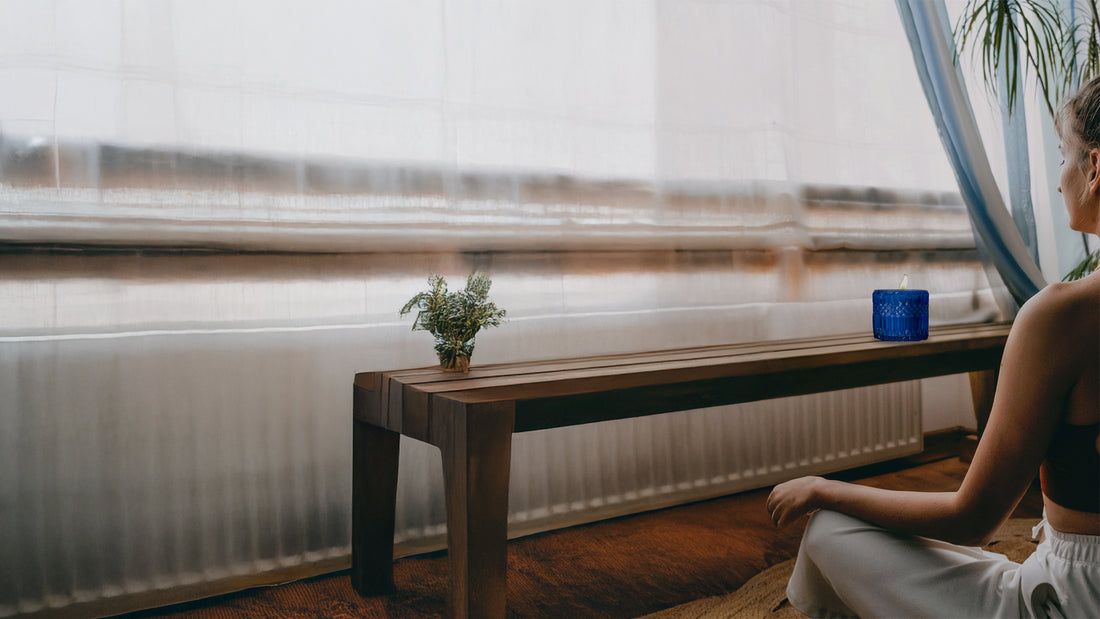 Person sitting near a window with a lit blue candle on a wooden bench, creating a calm and peaceful atmosphere.