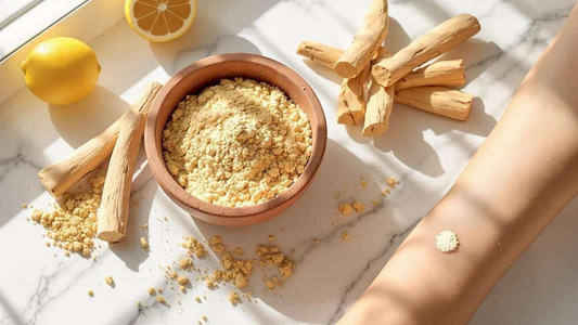 A bowl of yellow powder beside lemon slices and sticks of wood, with a small amount of powder on a bare arm against a marble surface.