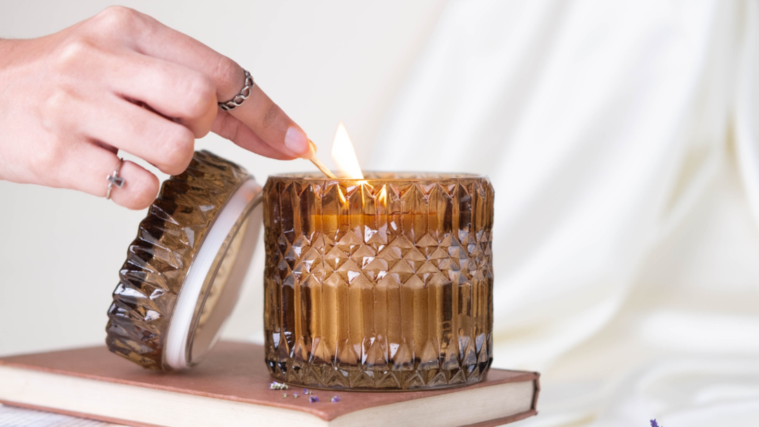 A hand lights a candle with a match, set on a stack of books, with a softly blurred background. The candle is in a textured amber glass holder.